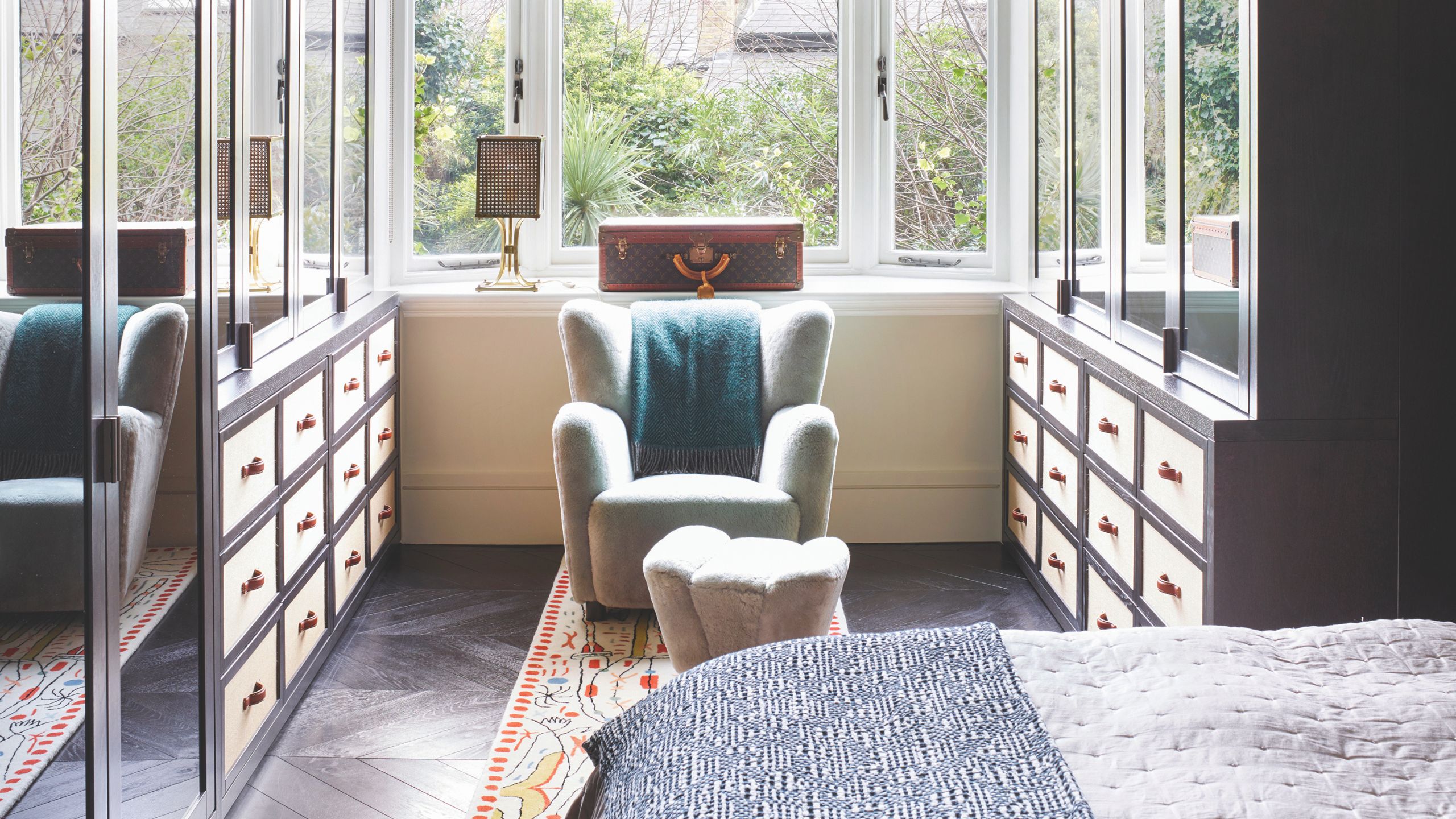 Bedroom with adjoining dressing room with mirrored closets and blue armchair, with large windows and leafy garden seen beyond.