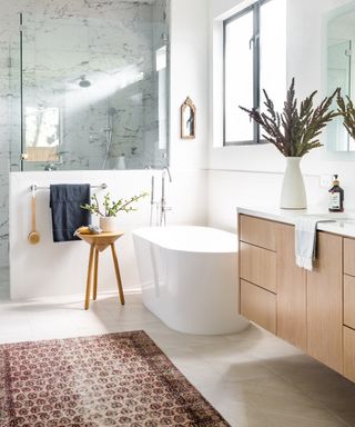 Bathroom with freestanding bathtub, wood fronted vanity, walk-in shower with marble tile and white tile floor with red patterned rug