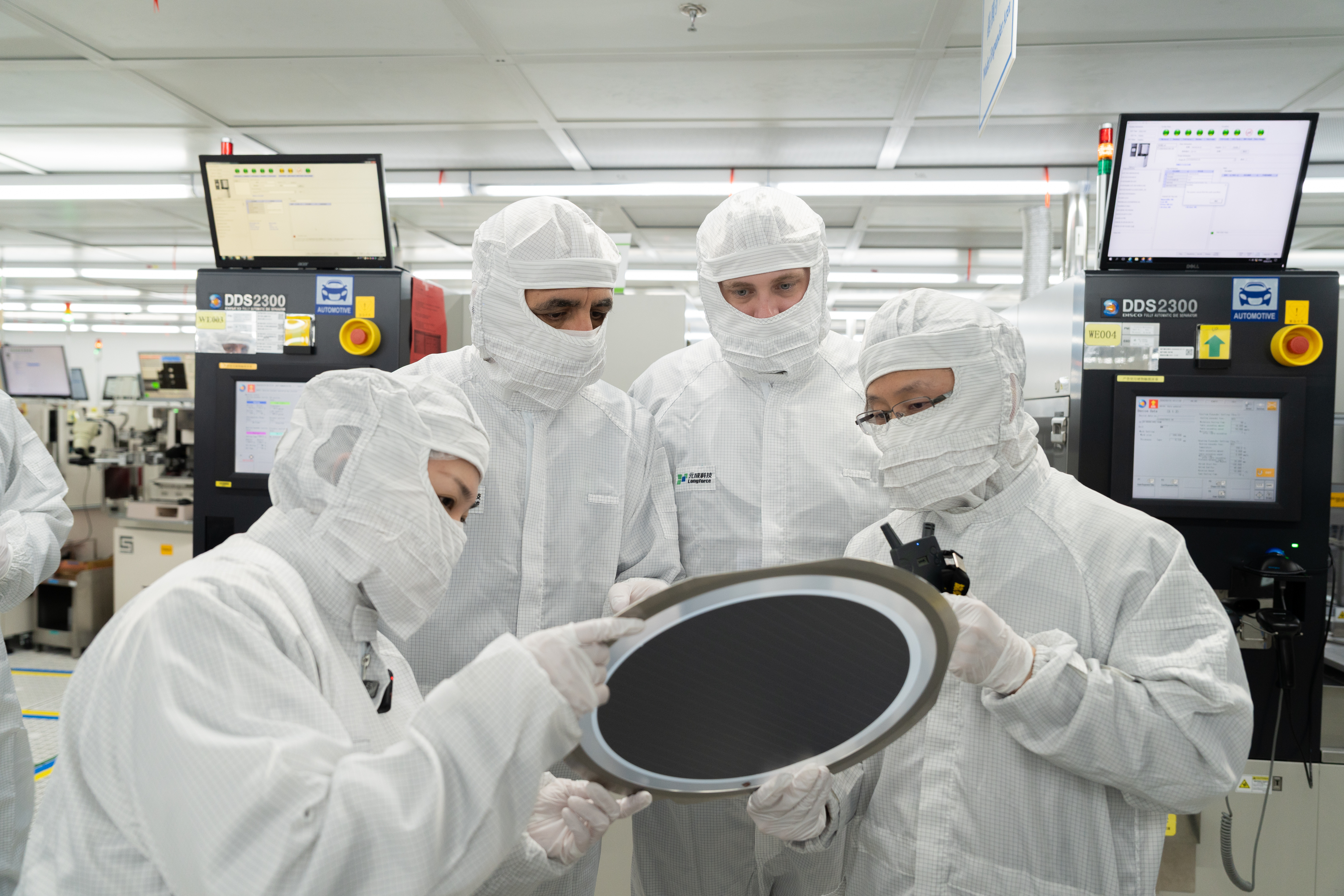Four people in full white cleanroom suits and masks gather around to inspect a large, dark semiconductor wafer in a high-tech manufacturing facility.