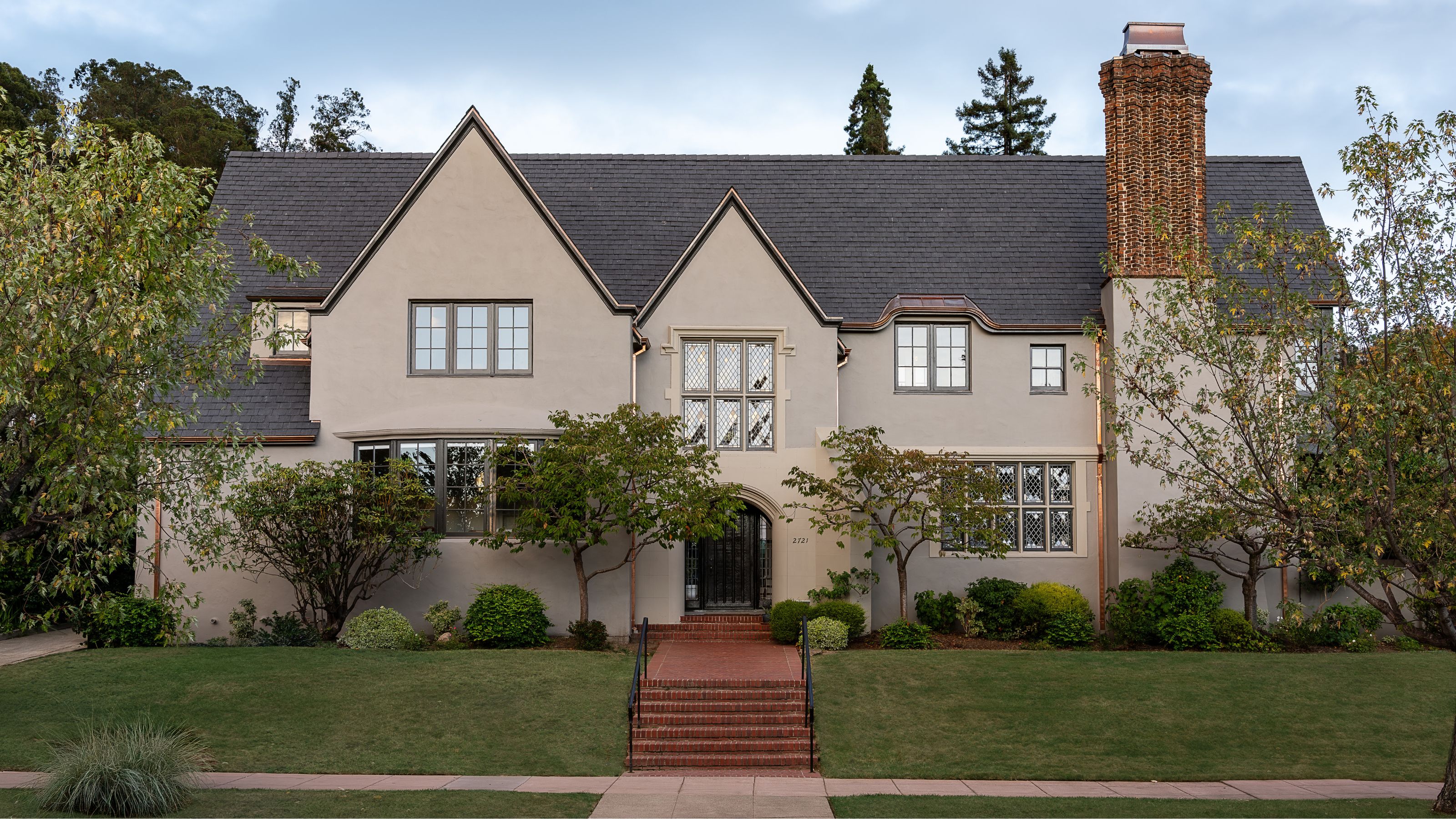 a neutral grand home with a large chimney, grass frontage with steps up to the front door and a series of small trees planted