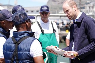 Prince William holding a fish as he speaks with fishermen at a harbor