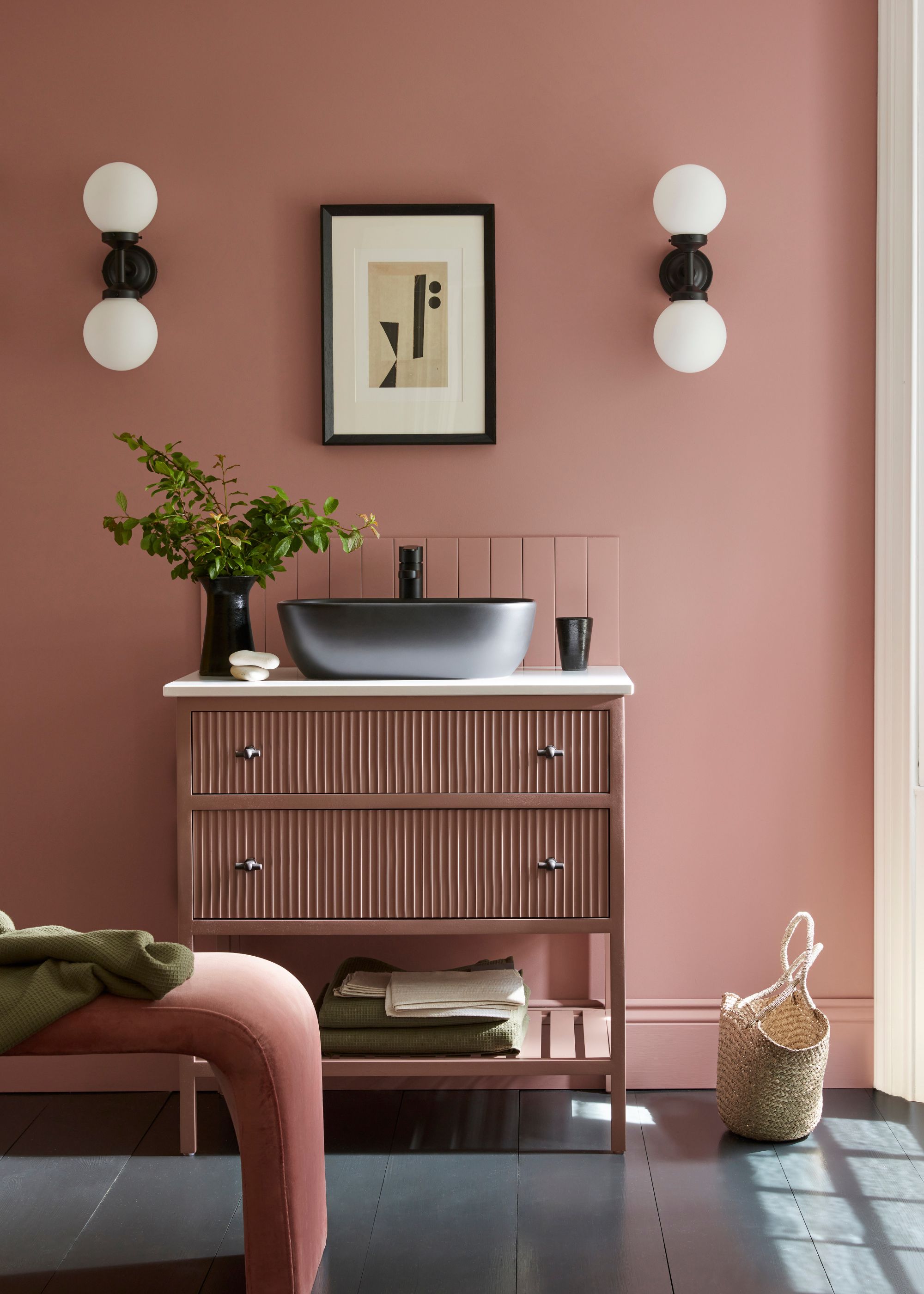 Bathroom with pink walls and pink vanity unit, round wall sconces, and framed artwork above the sink.