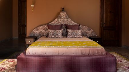 bedroom with orange walls, a patterned sculptural headboard with matching throw cushions, light pink sheets, a bright yellow throw, checked purple bedframe and vase, and patterned rug