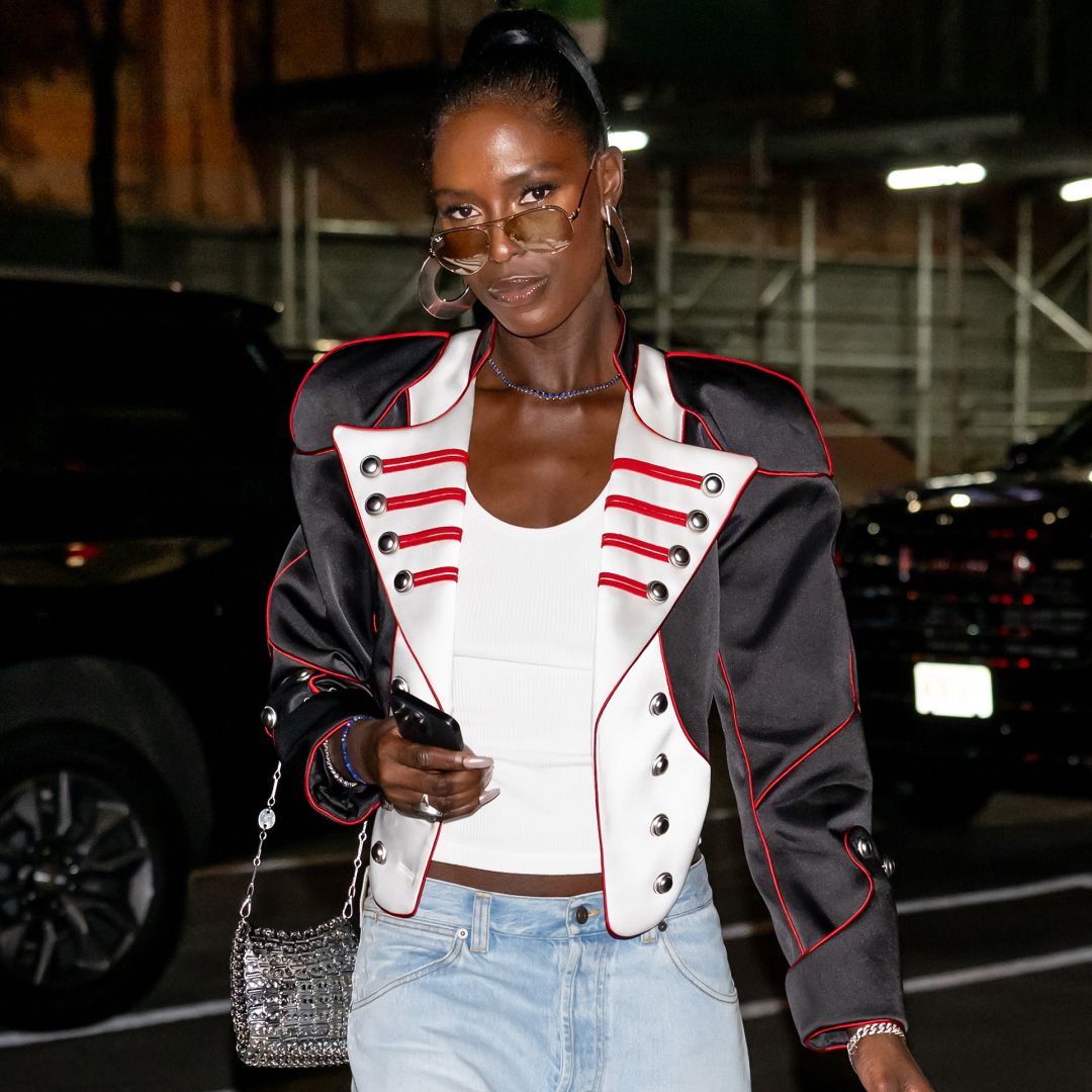 Jodie Turner Smith wearing a high ponytail and a white tank top, She has a jacket over the tank top and is wearing a pair of aviator sunglasses.
