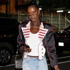 Jodie Turner Smith wearing a high ponytail and a white tank top, She has a jacket over the tank top and is wearing a pair of aviator sunglasses.