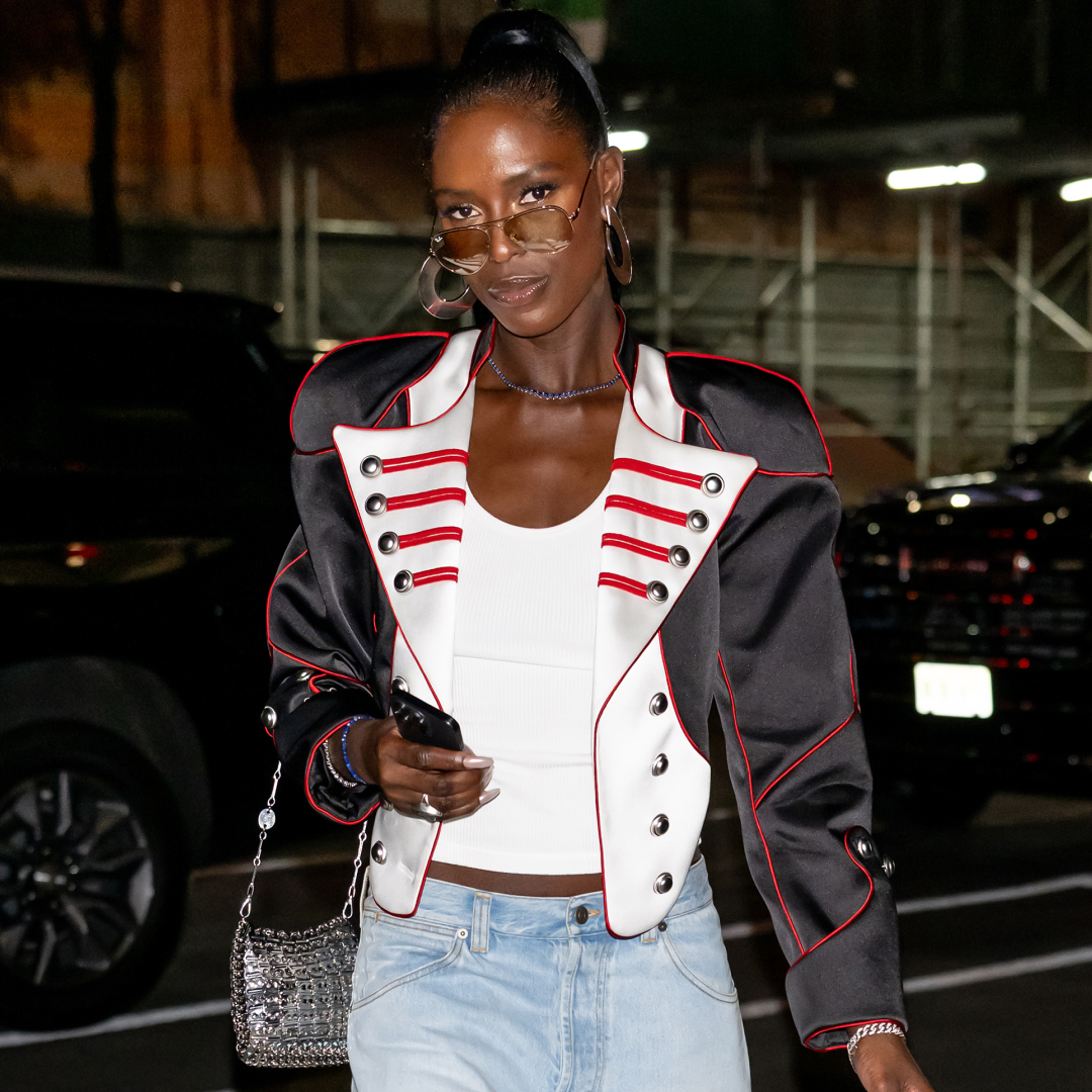 Jodie Turner Smith wearing a high ponytail and a white tank top, She has a jacket over the tank top and is wearing a pair of aviator sunglasses.