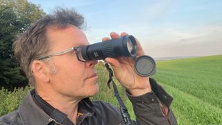 A man with glasses holding a monocular up to his right eye with a green field and blue sky in the background. 