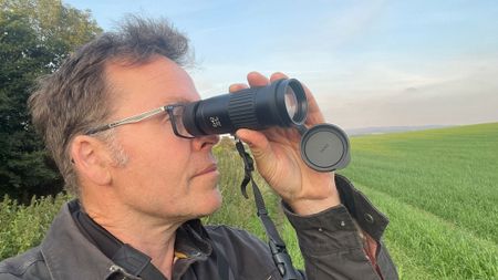 A man with glasses holding a monocular up to his right eye with a green field and blue sky in the background. 