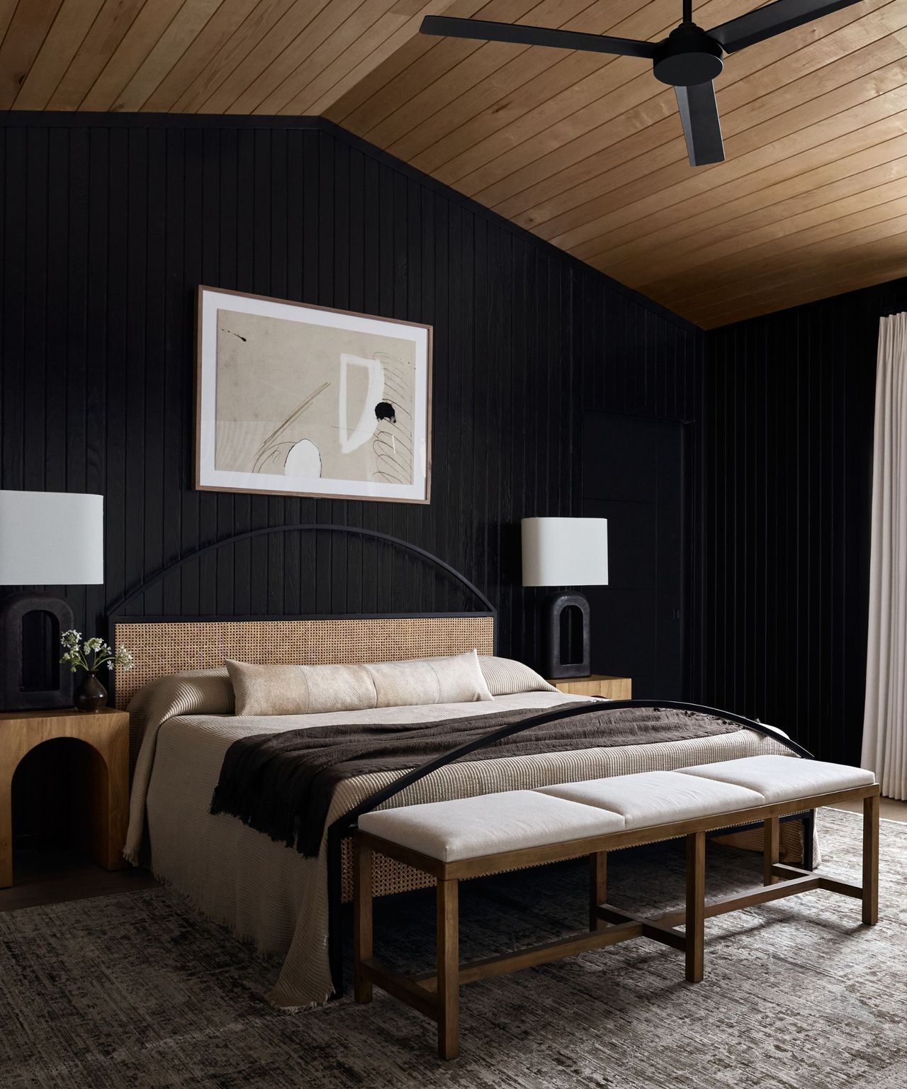 A coastal modern bedroom with black walls, a wood-paneled ceiling and a rattan bed frame