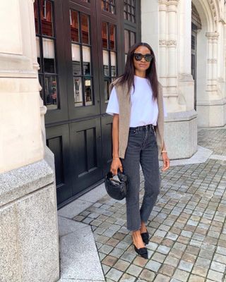 Influencer Lorna Humphrey poses on a sidewalk in London wearing oversize black sunglasses, a white t-shirt, neutral sweater over the shoulders, faded gray jeans, a black mini Bottega Veneta bag, and black suede mule heels.