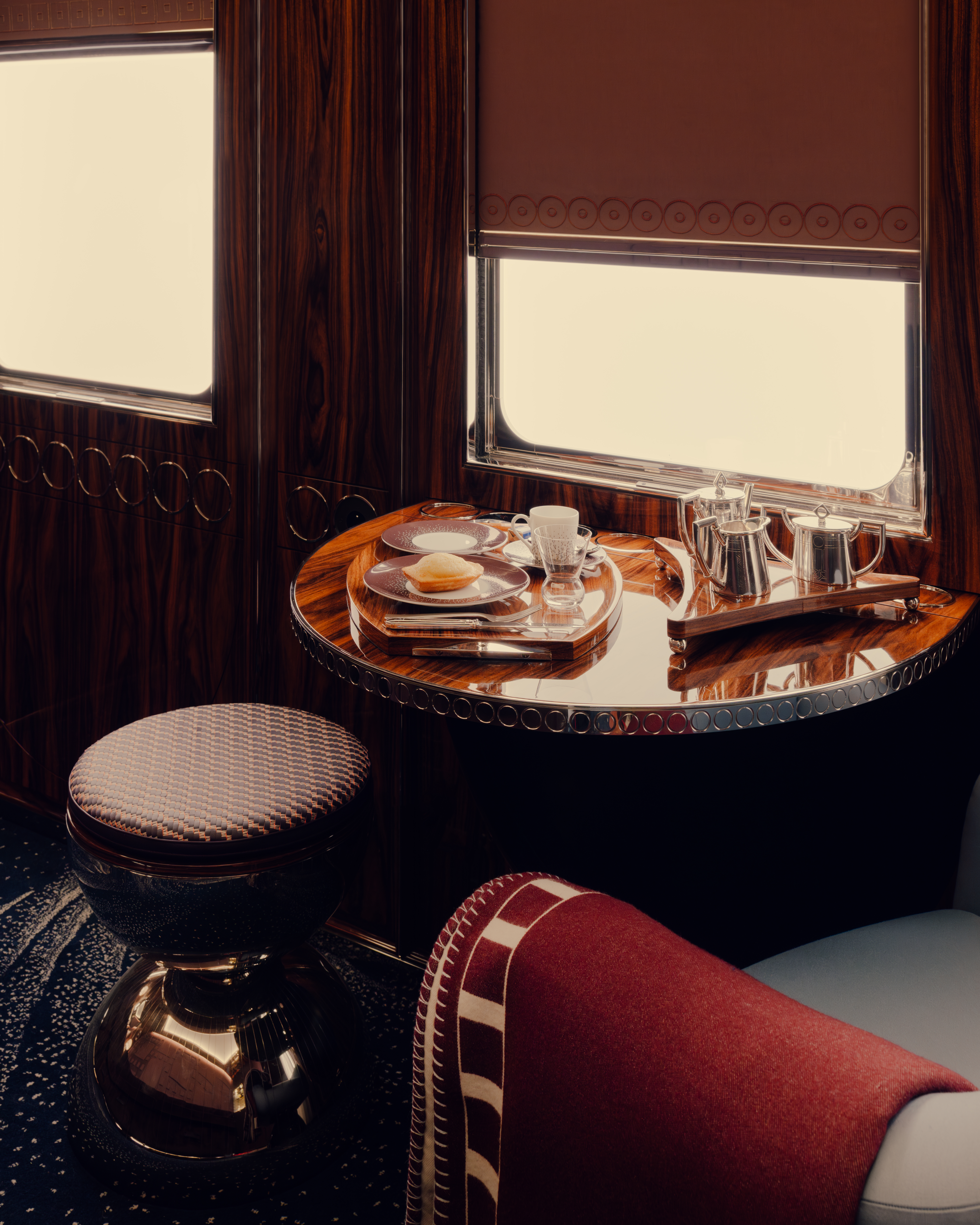 A window seat in a luxury train carriage, featuring sleek breakfast tableware, lacquered marquetry, and upholstered stools with checkered motifs in red and blue.