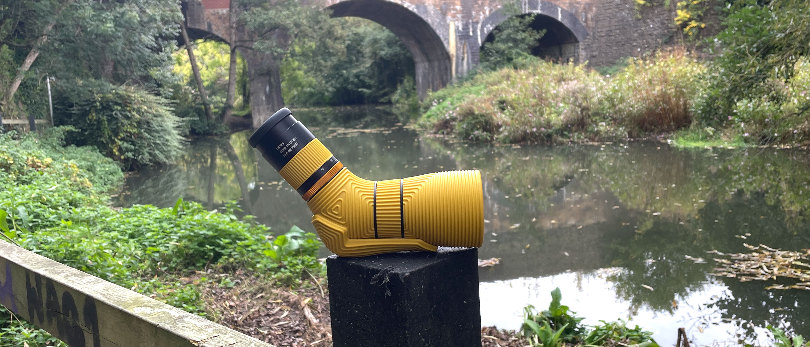 The Nocs Provisions Lite View spotting scope in yellow next to a river with a bridge in the background.