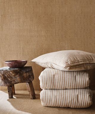 Striped bedding folded up on the floor next to a small wooden stool, a bowl on top. Orange/brown wallpaper behind.