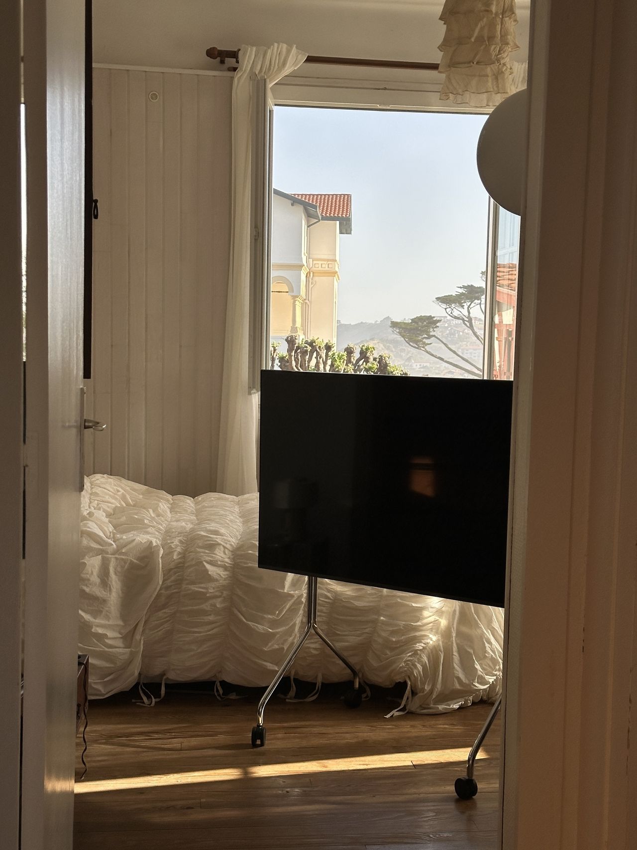 Image of a TV on a silver rolling tv stand in a white bedroom with a bed that has white, ruched bedding.
