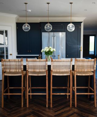 navy and white kitchen with dark wood floor, rattan and wood bar stools at kitchen island, integrated refrigerator, glass pendant lights