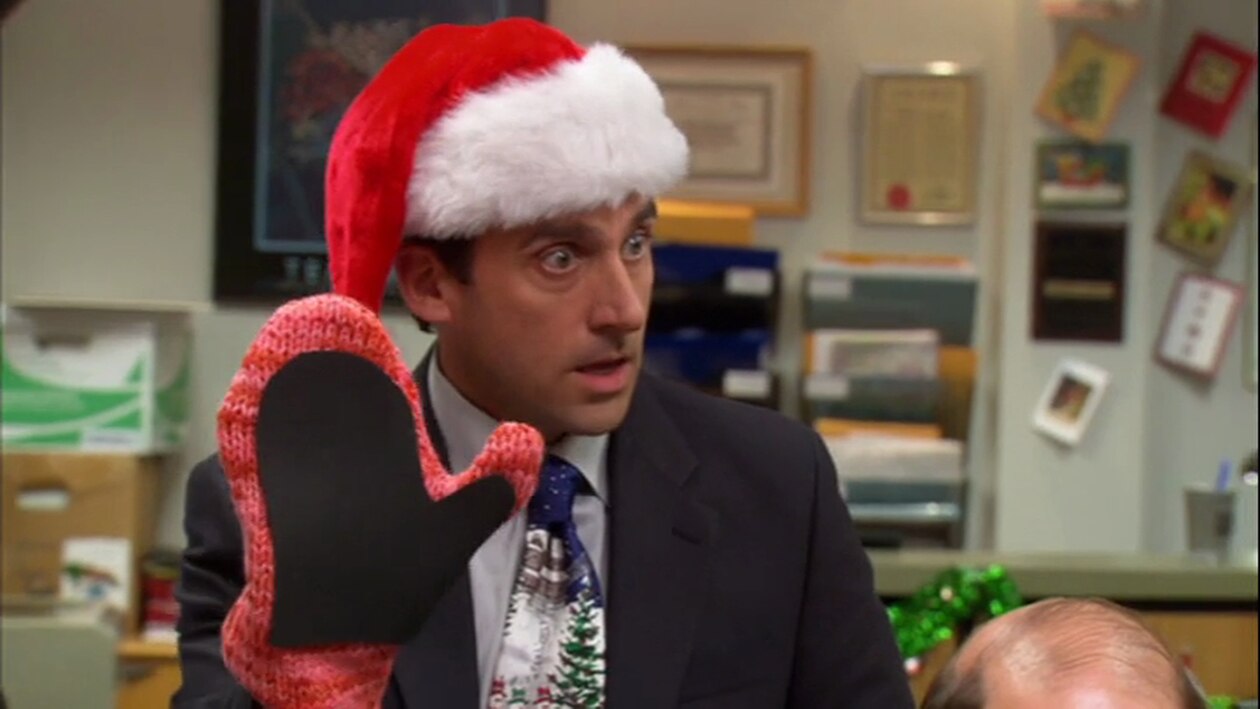 In a scene from The Office, Michael Scott wears a Santa hat and a festive tie while holding up a large red and black knitted mitten in an office setting.
