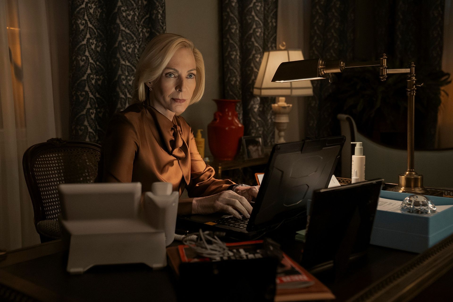 Allison Janney as Grace Penn, wearing an orange blouse while working at an office desk, in 'The Diplomat' season 4.
