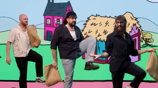 Three men traipsing in front of a fantastical cartoon land on a large screen behind them.