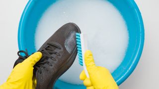Hands in rubber protective gloves holding black sport shoe and brush above blue basin