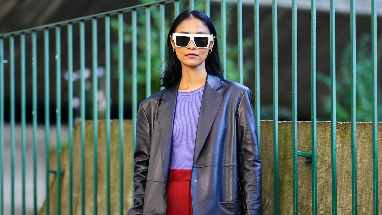 Pornwika Spiecker wears gold earrings, white sunglasses, a purple ribbed t-shirt, a black shiny leather long blazer jacket, high waist red large pants, a burgundy shiny leather large handbag, black and white leather pointed pumps heels shoes, outside the Stella McCartney show, during Paris Fashion Week - Womenswear Spring Summer 2022, on October 04, 2021 in Paris, France.