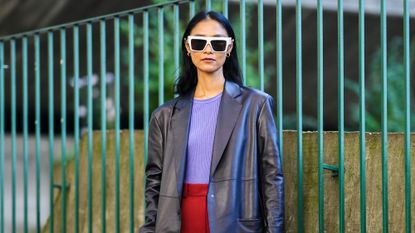 Pornwika Spiecker wears gold earrings, white sunglasses, a purple ribbed t-shirt, a black shiny leather long blazer jacket, high waist red large pants, a burgundy shiny leather large handbag, black and white leather pointed pumps heels shoes, outside the Stella McCartney show, during Paris Fashion Week - Womenswear Spring Summer 2022, on October 04, 2021 in Paris, France.