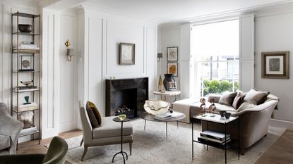 A white living room with neutral furniture, large windows, white panelled walls, a patterned pale Persian-style rug, and a black marble fireplace in the center of the back wall. To the left of the fireplace is a tall shelving unit with books and decorations.