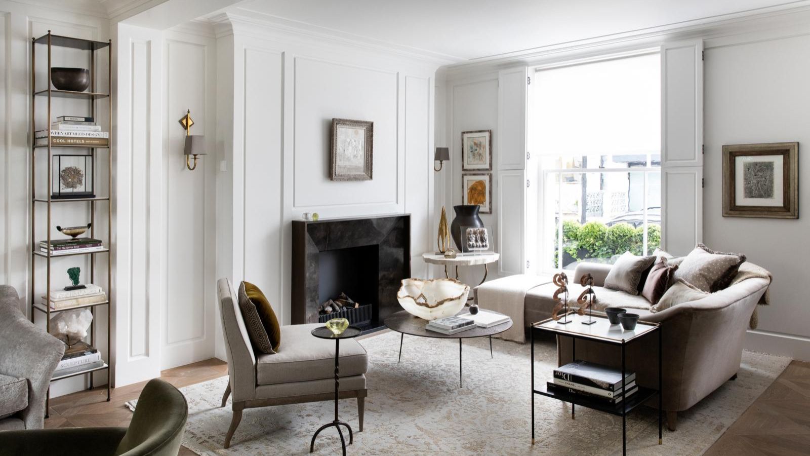 A white living room with neutral furniture, large windows, white panelled walls, a patterned pale Persian-style rug, and a black marble fireplace in the center of the back wall. To the left of the fireplace is a tall shelving unit with books and decorations.