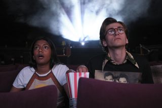 two teenagers (charithra chandran and sebastian croft) have shocked faces while watching a movie, with the projector shining behind them, in a still from 'how to date billy walsh'