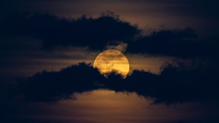 A yellow full moon is pictured glowing in a dark night sky between two worm-like bands of dark clouds.