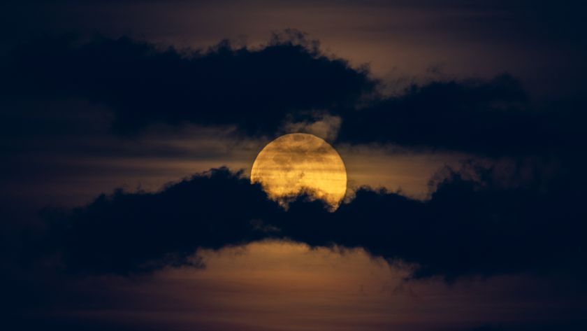 A yellow full moon is pictured glowing in a dark night sky between two worm-like bands of dark clouds.