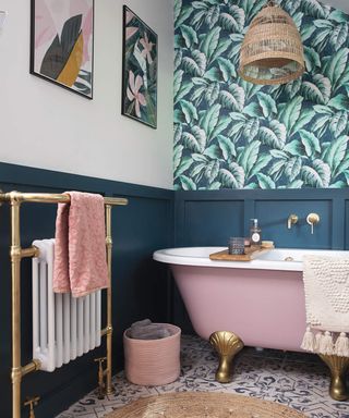 A bathroom with palm print wallpaper, blue wall paneling, pink bathtub, jute lampshade and jute rug