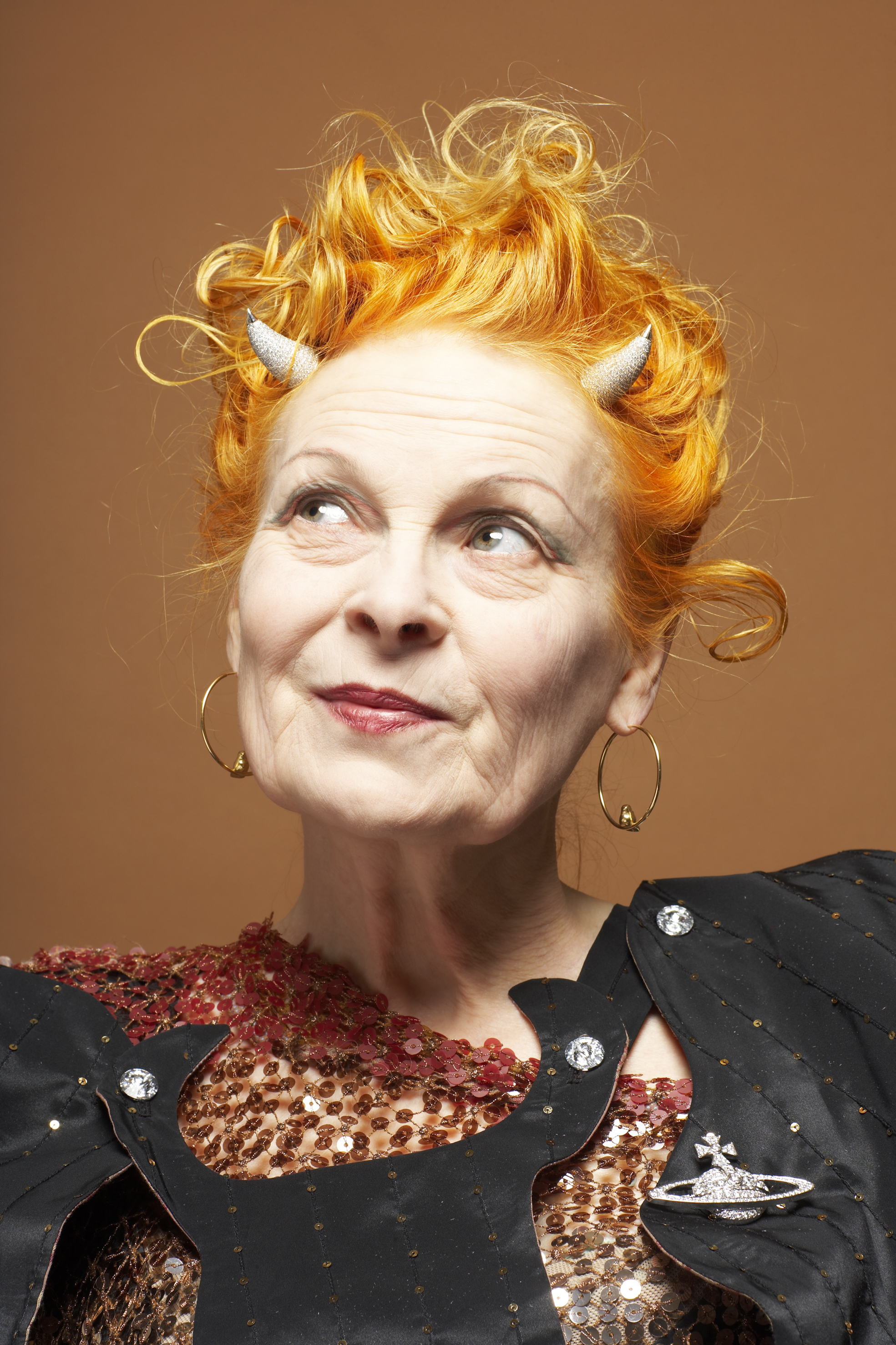 Vivienne Westwood looks upward with a mischievous expression, her wild orange hair adorned with two small silver horns, wearing a sequinned black jacket and her signature Saturn brooch.