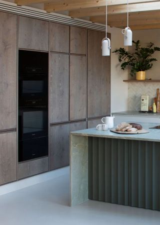 A bright kitchen island with bread cut up on a chopping board. The island has unique paneling cuts on one side, and on the other side, the countertop has been placed onto the side, meeting the floor. Next to the island are a few large cabinets and an oven.