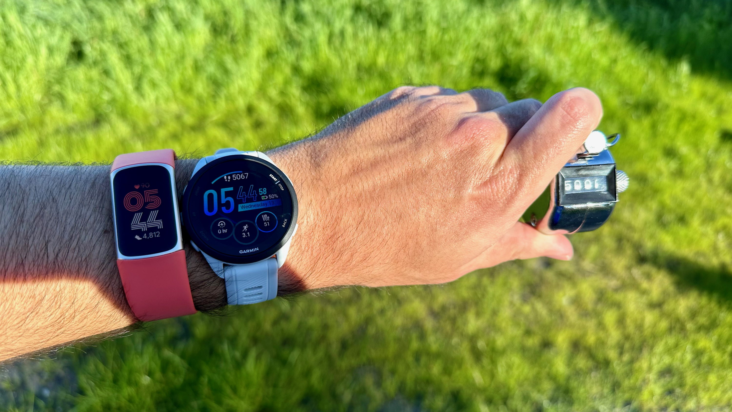 A pedometer held in hand with the number 5,000, a Garmin Forerunner 165 (5,062 steps), and a Fitbit Charge 6 (4,812 steps).