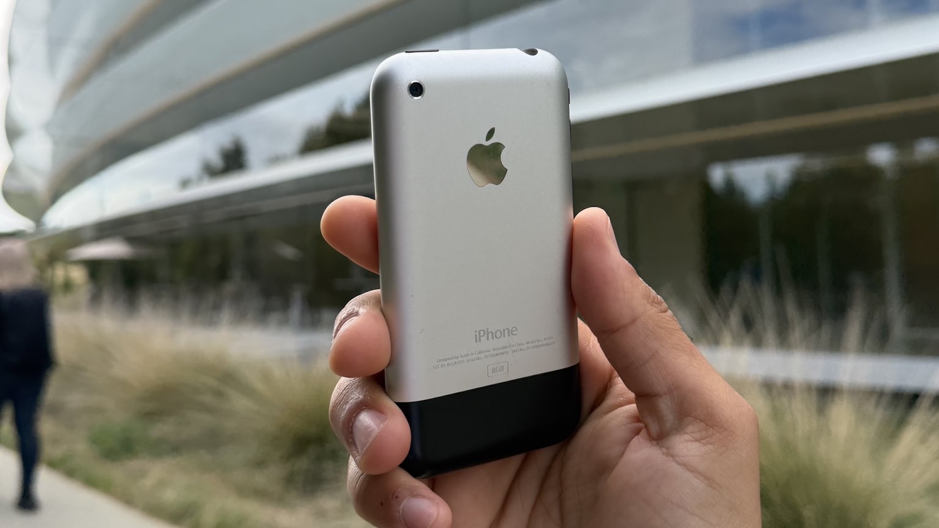 A hand holding up the original iPhone outside Apple Park