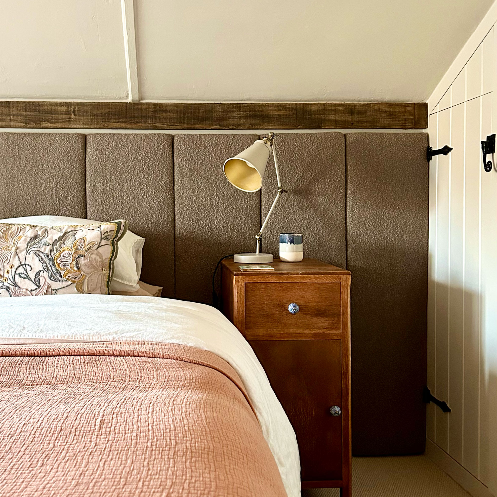 Country eaves bedroom with wide upholstered panelled headboard, wooden bedside table, lamp and pink throw