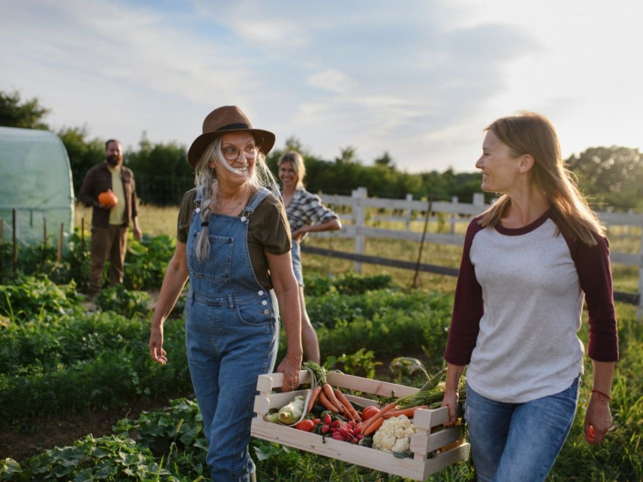 An older woman and a younger woman smile at each other while carrying a crate of vegetables through a garden