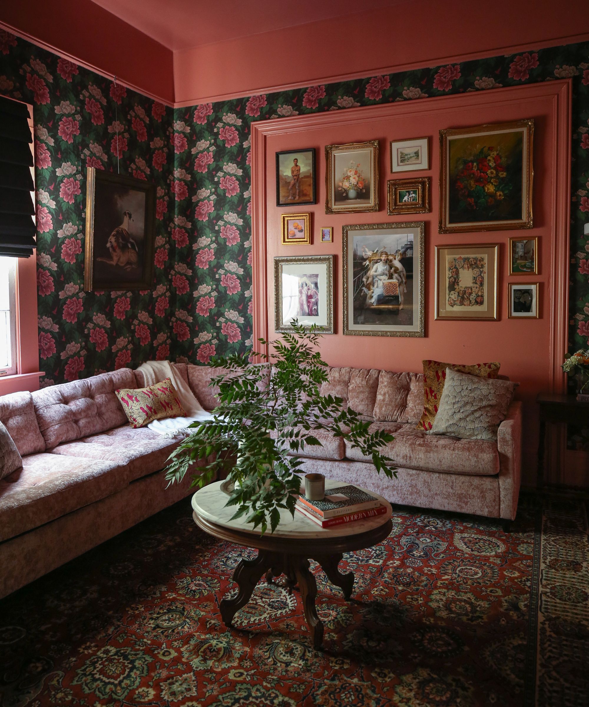 A living room with floral wallpaper, a gallery wall, a dusky pink L-shaped couch, and a marble table with a plant and books on top