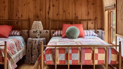 Rustic twin bedroom with wood paneling, sloped ceiling, an A-frame window overlooking trees, and bright patchwork quilts