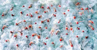 Top-down image of tens of swimmers with red swimming caps creating loads of splashes in the water 