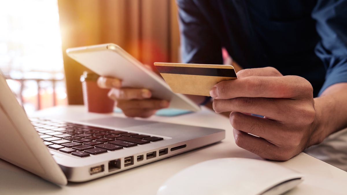 A photo of a person holding a bank card in one hand and their phone in the other, sitting at a laptop and making a purchase online