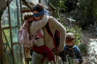 sandra bullock wearing a mask covering her eyes and walking with two children in a wooded trail in birdbox