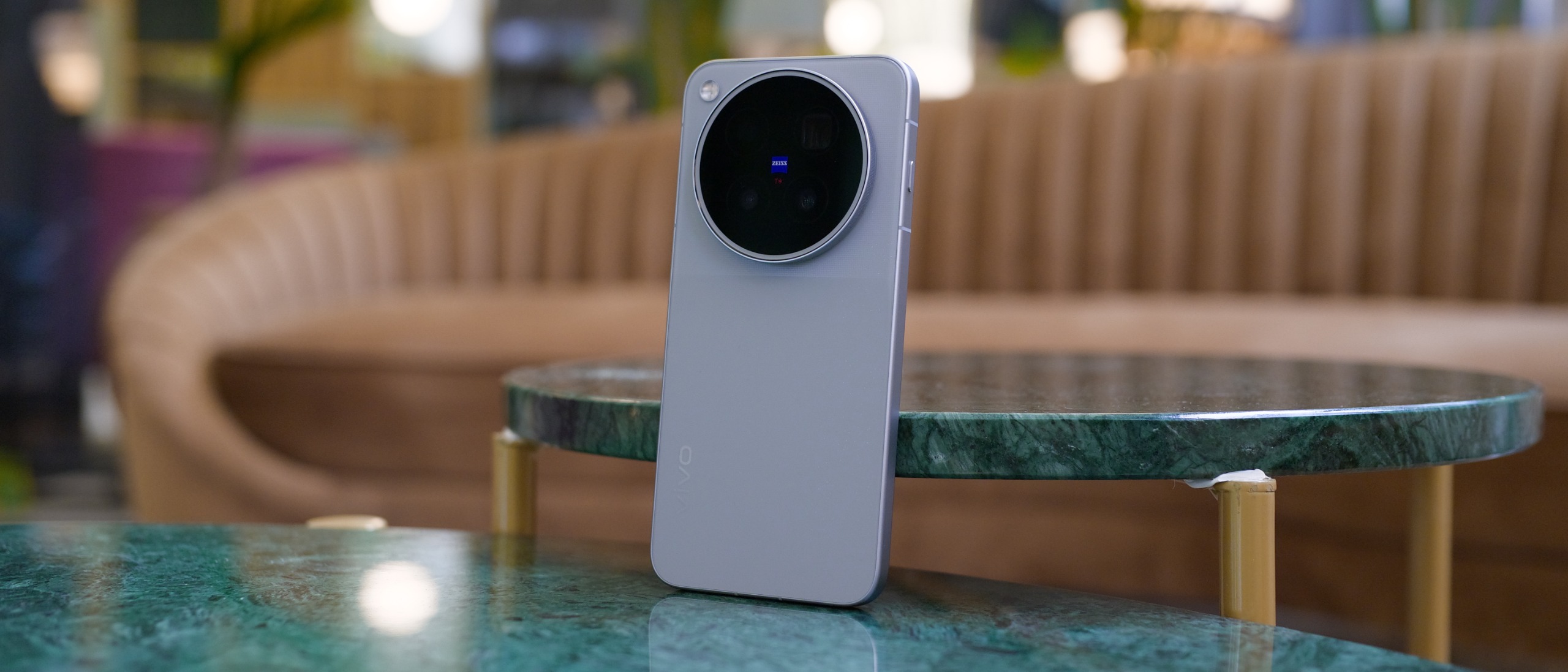 Vivo X500 Pro Max standing upright on a marble table, photographed from the rear with a blurred seating area in the background.