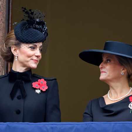 Kate Middleton wearing a black hat and coat turning to the right and smiling at Duchess Sophie, also wearing a black hat and coat with a red poppy brooch