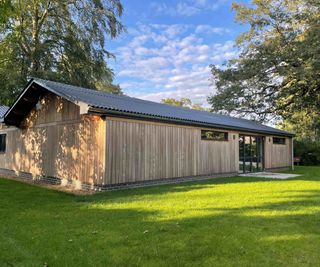 A large single storey outbuilding, set in a lawned garden with mature trees in the background. The building features bifold doors, three windows, two pitched roofs, and is clad in Vastern Timber's Brimstone modified timber cladding.