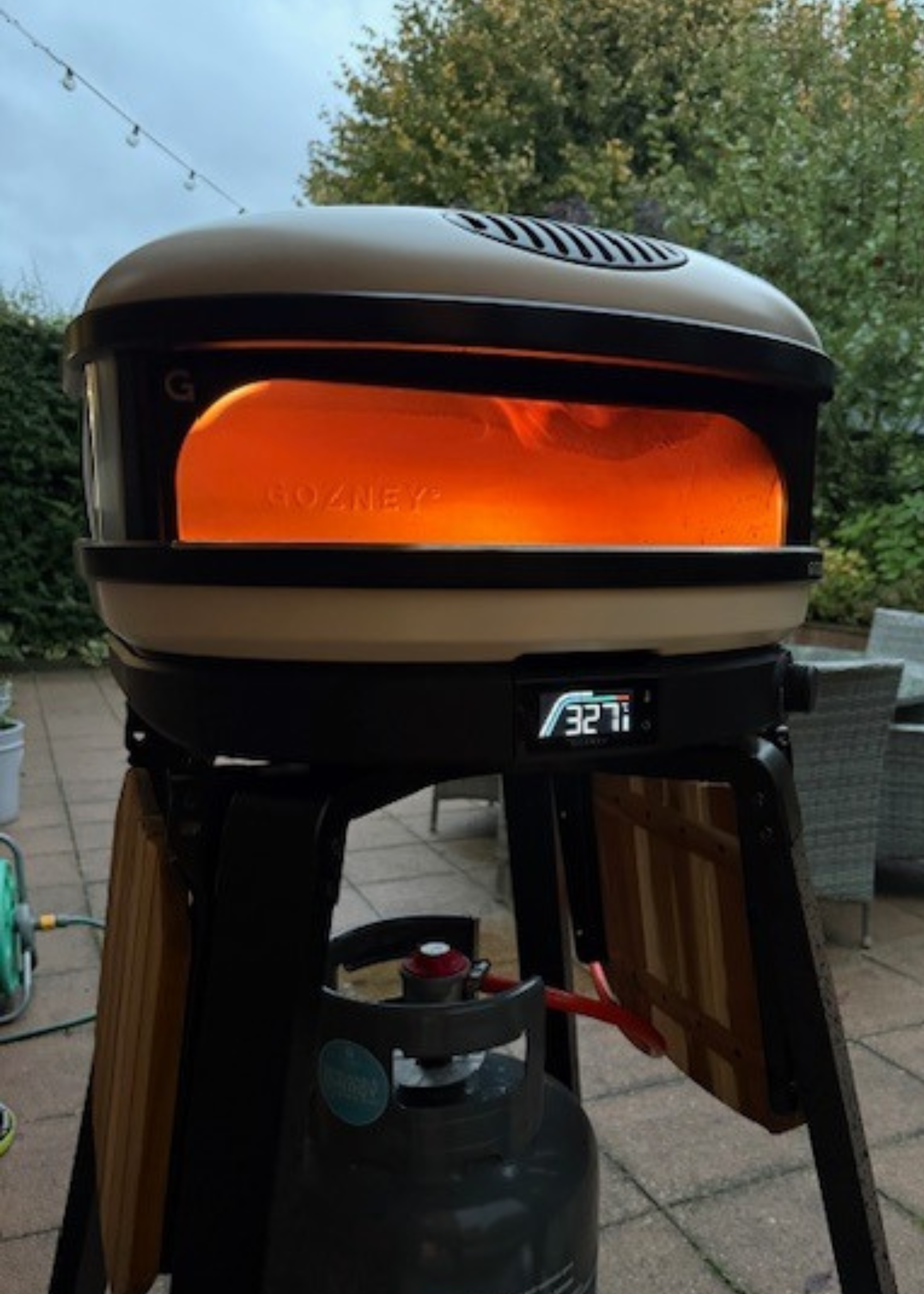 The Gozney Arc XL pizza oven heating up with the flame in view and a garden going into early evening darkness in the background behind