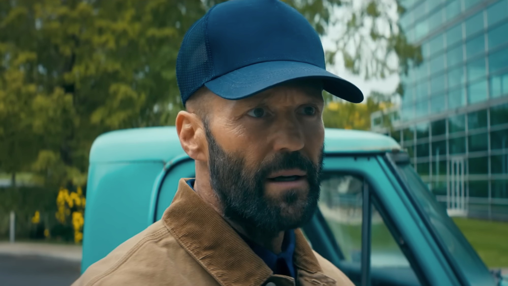 Jason Statham with a beard, wearing a blue hat and brown jacket in The Beekeeper.