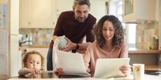 A mother and father look at a computer at the kitchen table while their child looking on.