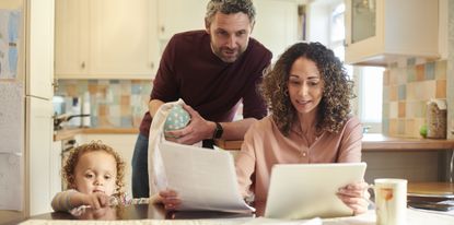 A mother and father look at a computer at the kitchen table while their child looking on.