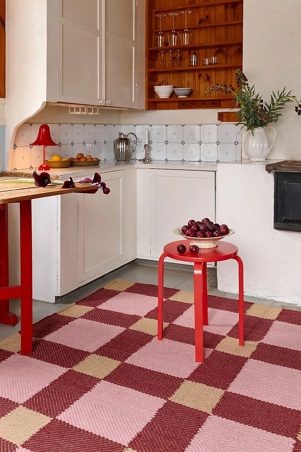 Image of a pink plaid rug in a white, modern farmhouse kitchen. There is a red stool on the rug that is holding a bowl full of cherries